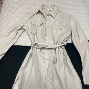 Beige läder kappa från H&M - Snygg beige läder kappa från H&M. Kappan har långa ärmar, knappar framtill och ett bälte i midjan för en smickrande passform. Perfekt för en stilren look.