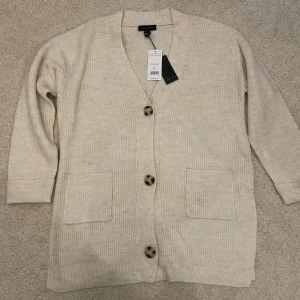 Beige ribbad/stickad kofta från New Look - Helt ny och oanvänd med prislappen kvar. Säljer en stilren beige kofta från Zara med ribbad struktur och stora knappar framtill. Koftan har långa ärmar och praktiska fickor. Slitsar på båda sidorna. Skriv för mer information 🩷