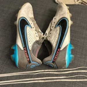 Nike Tiempo fotbollsskor - Säljer ett par Nike Tiempo fotbollsskor i vitt och blått med svarta detaljer. Skorna har snörning och är designade för optimal prestanda på planen. Perfekta för fotbollsspelare som söker komfort och stil.