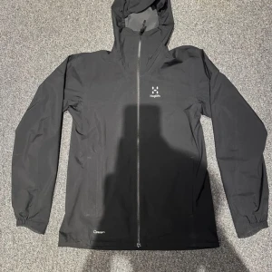  Haglöfs jacka  - Snygg och funktionell svart vindjacka från Haglöfs med dragkedja och huva. Perfekt för blåsiga dagar och utomhusaktiviteter. Jackan har långa ärmar och en stilren design.