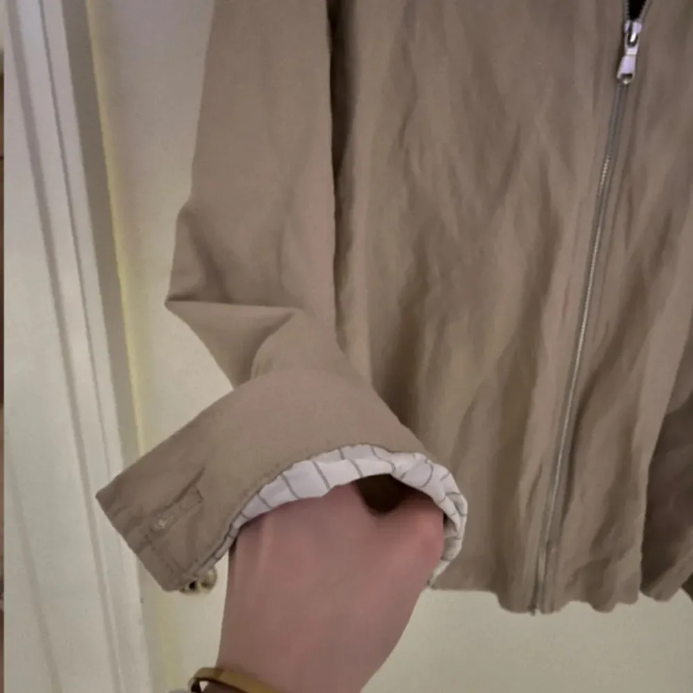 Snygg beige jacka från J.Lindeberg med dragkedja framtill. Jackan har en klassisk krage och långa ärmar med uppvikta ärmslut. Perfekt för en stilren look.. Neuletakit & Villapaidat.