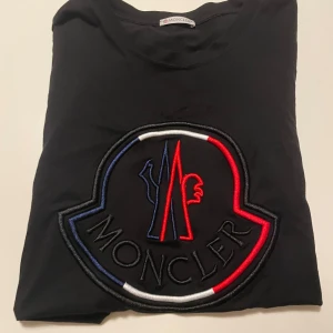 Svart t-shirt från Moncler - Snygg svart t-shirt från Moncler med ett stort broderat märke i rött, blått och vitt på framsidan. T-shirten har korta ärmar och är perfekt för en stilren look.
