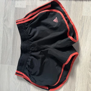 Svarta träningsshorts från Adidas!😍 - Säljer ett par svarta träningsshorts från Adidas med röda detaljer. Shortsen har en elastisk midja och är perfekta för löpning eller annan träning. Adidas-loggan syns tydligt på framsidan. Sköna och funktionella för aktiva dagar!😻 Shotsen kommer inte till användning, så de säljer jag nu🥰