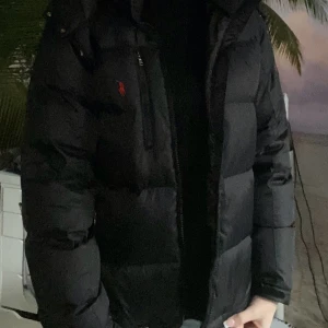 Svart dunjacka från Polo Ralph Lauren - Snygg svart dunjacka från Polo Ralph Lauren med röd broderad logga på bröstet. Jackan har en dragkedja framtill och är perfekt för kyligare väder. Den är långärmad och har en klassisk design som passar till mycket.