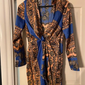 Mönstrad klänning från Missguided - Snygg mönstrad klänning från Missguided i blå och orange med långa ärmar och omlott-design. Perfekt för en färgglad look.