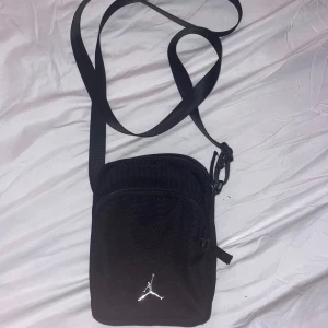 Svart axelväska från Jordan - Snygg svart axelväska från Jordan med justerbar rem och dragkedja. Perfekt för att bära med sig det nödvändigaste. Väskan har en stilren design med det ikoniska Jordan-logotypen i silver på framsidan.