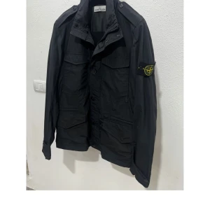 Svart jacka från Stone Island - Snygg svart jacka från Stone Island med flera fickor och knappar framtill. Jackan har en klassisk design med en broderad logga på ärmen. Perfekt för en stilren look. Säljer jackan då den precis har blivit lite för liten för mig. Jackan är äkta och storlek M 