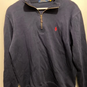 Mörkblå tröja med dragkedja från Ralph Lauren - Snygg mörkblå tröja från Ralph Lauren med en halv dragkedja och det klassiska röda logotypbroderiet på bröstet. Perfekt för en stilren och avslappnad look.