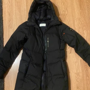 Svart dunjacka från Sail Racing - Snygg svart dunjacka från Sail Racing med huva och dragkedja. Perfekt för kalla dagar med sin varma och vadderade design. Jackan har en stilren logga på bröstet och är tillverkad för att klara tuffa väderförhållanden. Använd ett par gånger men ser helt ny ut. 