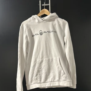 Vit hoodie från Sail Racing - Säljer en stilren vit hoodie från Sail Racing med svart logotyp på bröstet. Tröjan har en klassisk känguruficka och en bekväm huva. Perfekt för en avslappnad look.