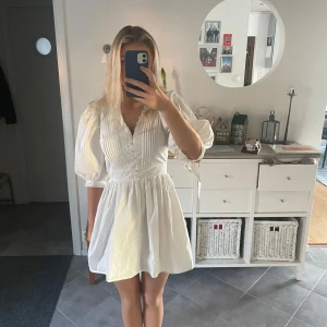 Vit kortklänning med knappar - Säljer en söt vit kortklänning med puffiga trekvartsärmar och knappar framtill. Klänningen har en v-ringad design och ett knytband i midjan för en fin passform. Perfekt för sommarens alla tillfällen!