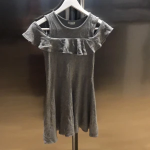 Grå klänning med volangdetalj - Säljer en söt grå klänning med volangdetalj runt axlarna. Klänningen har en offshoulder-design och är tillverkad i ett mjukt material. Perfekt för en avslappnad stil.