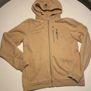 Beige hoodie från Sail Racing - Snygg beige hoodie från Sail Racing med dragkedja och justerbar huva. Perfekt för en avslappnad stil. Den har en bröstficka med dragkedja och logotyp på bröstet. Nypris 1000kr