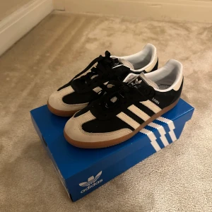 Adidas Samba sneakers - Snygga Adidas Samba sneakers i svart och vit med klassiska tre ränder på sidan. Skorna har en gummisula och snörning för en bekväm passform. Perfekta för en stilren och sportig look.