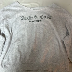 Grå sweatshirt med tryck - Säljer en grå sweatshirt med texten 'Mind & Body Movement' tryckt på framsidan. Tröjan har långa ärmar och en avslappnad passform, perfekt för en chill dag. Mjuk och bekväm att bära.