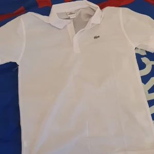 Vit pikétröja från Lacoste - Säljer en klassisk vit pikétröja från Lacoste med den ikoniska krokodilloggan på bröstet. Tröjan har korta ärmar och en stilren krage med knappar. Perfekt för en avslappnad och stilren look.