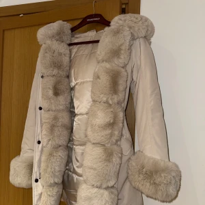 Beige parkajacka med fuskpäls - Snygg beige parkajacka med mjuk fuskpäls på huva, ärmslut och längs med dragkedjan. Jackan har en justerbar midja med knytband och praktiska fickor. Perfekt för kyliga dagar.säljes pga inte kommer till användning 