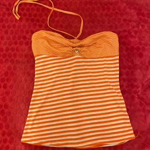 Orange- och vitrandig tubtopp - Snygg orange- och vitrandig tubtopp med dekorativa stenar framtill. Toppen har en knytbar halterneck och är perfekt för sommardagar. Den är gjord i ett mjukt och bekvämt material.