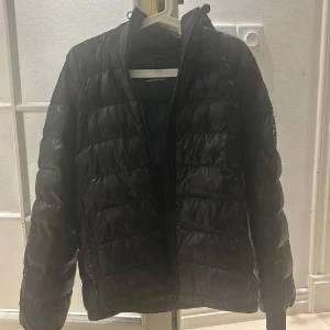 Svart dunjacka från Peak Performance - Säljer en svart dunjacka från Peak Performance. Jackan har en stilren design med dragkedja och är perfekt för kyligare väder. Den är långärmad och har en praktisk huva för extra skydd mot vind och kyla.