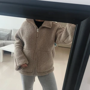 Beige teddyjacka med dragkedja - Mysig beige teddyjacka med dragkedja framtill och stora fickor. Perfekt för kyliga dagar med sin fluffiga och varma design. Jackan har en avslappnad passform och långa ärmar. Det är XS men väldigt stor för sin storlek så passar även S