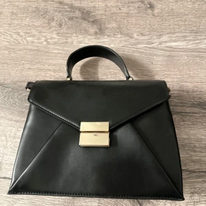 Svart handväska med gulddetaljer - Elegant svart handväska i skinn med guldfärgad låsdetalj framtill. Väskan har ett praktiskt handtag och en dragkedja på baksidan för extra förvaring. Perfekt för en stilren look.