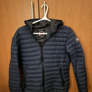 Mörkblå pufferjacka från Colmar - Säljer en riktigt snygg mörkblå pufferjacka från Colmar. Jackan har en dragkedja framtill och är quiltad för extra värme. Den har en huva och långa ärmar, perfekt för kyligare dagar.