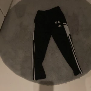Svarta träningsbyxor från EA - Säljer ett par svarta träningsbyxor från EA med vita ränder längs sidorna. Byxorna har en tight passform och är perfekta för träning. De har en elastisk midja för extra komfort.