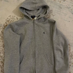 Grå hoodie från Polo Ralph Lauren - Säljer en klassisk grå hoodie från Polo Ralph Lauren med dragkedja och huva. Tröjan är i supefint skick men har tyvärr blivit för liten. Vid frågor eller ytterligare bilder är det bara att höra av sig! Pris kan diskuteras 