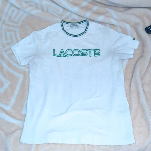 Vit t-shirt från Lacoste - Säljer en vit t-shirt från Lacoste med grönt tryck på bröstet. T-shirten har en klassisk rund halsringning med gröna detaljer. Perfekt för en avslappnad stil.