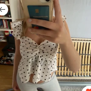 Vit prickig topp med volangdetaljer - Charmig vit topp med svarta prickar och volangdetaljer. Toppen har en knytning framtill och är ärmlös med breda axelband. Perfekt för en somrig look🥰🥰🥰pris kan diskuteras