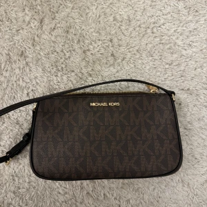 Brun axelväska från Michael Kors - Snygg brun axelväska från Michael Kors med monogrammönster och guldfärgad dragkedja. Väskan har en justerbar axelrem och är perfekt för att bära dina nödvändigheter med stil.  Säljer pågrund av att den en kommer till användning längre, den är ungefär ett halvår gammal! Vill du se kvitto skriv privat 😊