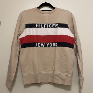 Beige sweatshirt från Tommy Hilfiger - Säljer en beige sweatshirt från Tommy Hilfiger med texten 'Hilfiger New York' i svart, vitt och rött. Tröjan har långa ärmar och ribbade muddar vid ärmslut och nederkant. Perfekt för en avslappnad stil!