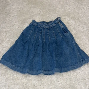 Blå jeanskjol - Säljer en söt blå jeanskjol med veckad design och elastisk midja. Perfekt för en avslappnad stil. Kjolen har en klassisk look och är lätt att matcha med olika toppar.