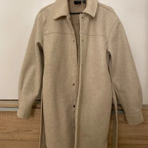 Beige kappa från Vero Moda - Snygg beige kappa från Vero Moda med knappar och bälte i midjan. Perfekt för höst och vår. Långärmad design med klassisk krage och stilren look. Använd några gånger men växt ur den!