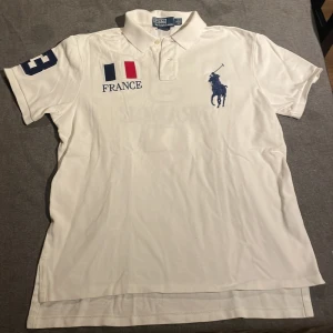 Vit pikétröja från Ralph Lauren - Säljer en vit pikétröja från Ralph Lauren med broderad logga och fransk flagga. Tröjan har korta ärmar och nummerdetaljer på ärmen och ryggen. Perfekt för en chief keef look lowkey.