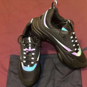 Dior B22 - Snygga svarta sneakers från Dior med coola iriserande detaljer i lila och blått. Skorna har snörning och en robust sula för extra komfort. Perfekta för att ge din outfit en lyxig touch.