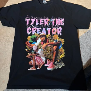 Tyler the Creator T-shirt - Svart T-shirt med tryck av Tyler, the Creator och texten 'Flower Boy'. Framsidan har en design med blommor och en lista av låttitlar på baksidan.