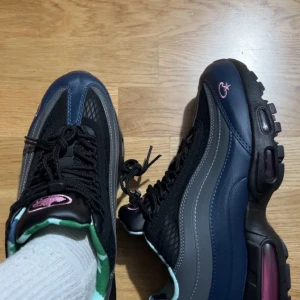 Corteiz X Nike Air Max 95 i svart och rosa - Snygga Nike Air Max sneakers i svart och blått med rosa detaljer. Skorna har en klassisk Air Max-sula och snörning framtill. Perfekta för en sportig look.