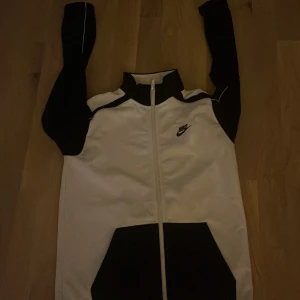 Vit och svart jacka från Nike användt 2 gånger  - Säljer en stilren vit och svart jacka från Nike med dragkedja och hög krage. Jackan har långa ärmar och ett klassiskt Nike-märke på bröstet. Perfekt för en sportig look! Är i jätte bra skick bara användt den 2 gånger 