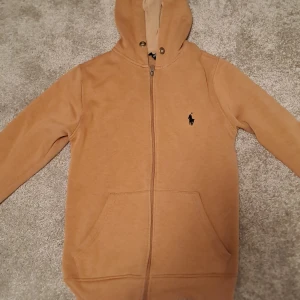 Beige hoodie från Ralph Lauren - Säljer en snygg beige hoodie från Ralph Lauren med dragkedja och klassisk logga på bröstet. Perfekt för en avslappnad stil. Den har långa ärmar och en praktisk huva.