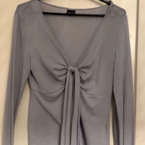 Grå långärmad topp från Gina Tricot - Snygg grå långärmad topp från Gina Tricot med knytning framtill. Perfekt för en stilren look. Toppen har en v-ringad design och är tillverkad i ett mjukt material.