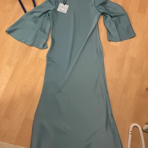 Blå långklänning med vida ärmar - Elegant grön långklänning med vida ärmar. Perfekt för en stilfull kväll eller speciellt tillfälle. Klänningen har en enkel och sofistikerad design som framhäver en vacker siluett.