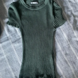 Grön ribbad topp från Zara - Snygg grön ribbad topp från Zara med korta ärmar. Perfekt för en stilren look. Toppen har en tight passform och är tillverkad i ett mjukt material som ger en bekväm känsla. Den är ganska genomskinlig 