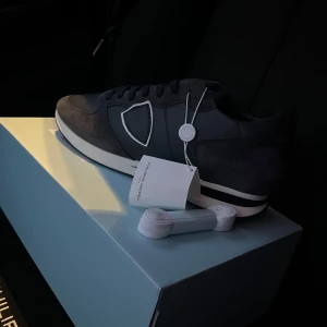 Mörkblå sneakers från Philippe Model - Stilrena mörkblå sneakers från Philippe Model med vita detaljer och snörning. Skorna har en klassisk design med en diskret logotyp på sidan. Perfekta för en avslappnad och trendig look.