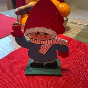 Dekorativ tomtefigur med röd luva - Charmig träfigur föreställande en tomte med röd luva och blå kläder. Tomten håller en ljushållare i handen, perfekt för juldekoration. Den har en vit och röd randig halsduk och står på en grön bas.