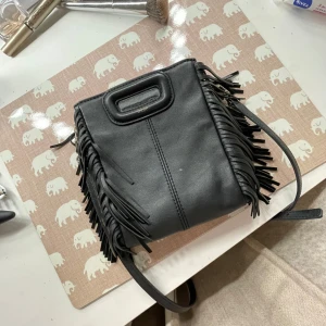 Svart axelväska med fransar från Maje - Snygg svart axelväska i skinn från Maje med coola fransdetaljer på sidorna. Både läderband och kedjeband ingår. Har haft den i ungefär ett år och är fortfarande i superbra skick, bara en liten vitt fläck! Pris kan diskuteras🩷