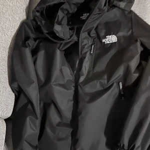 Svart vindjacka från The North Face - Säljer en svart vindjacka från The North Face med huva och dragkedja. Perfekt för blåsiga dagar och har en stilren design med logotyp på bröstet. Jackan är lätt och smidig att bära med sig.