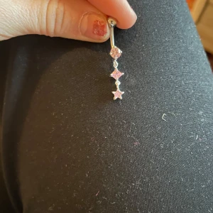 Silverfärgat navelpiercingsmycke med stjärnor - Snyggt navelpiercingsmycke i silverfärg med tre hängande stjärnor. Smycket har en glittrig design med rosa stenar som ger en extra touch av glamour. Perfekt för att lysa upp din stil!