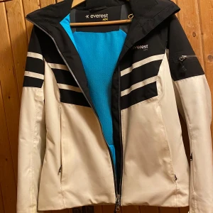 Skidjacka Everest - Snygg vit och svart jacka från Everest med dragkedja och huva. Jackan har svarta detaljer och ett blått innerfoder. Perfekt för kyligare väder. Strl 36. Köpt på stadium för 1799kr. 