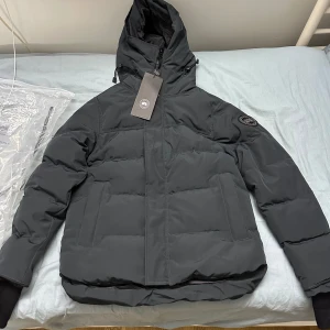 Svart pufferjacka med huva - Snygg svart canada gosse black label med huva och dragkedja. Perfekt för kalla dagar med sin vadderade design. Jackan har långa ärmar och en stilren look. Helt ny jacka oanvänt 